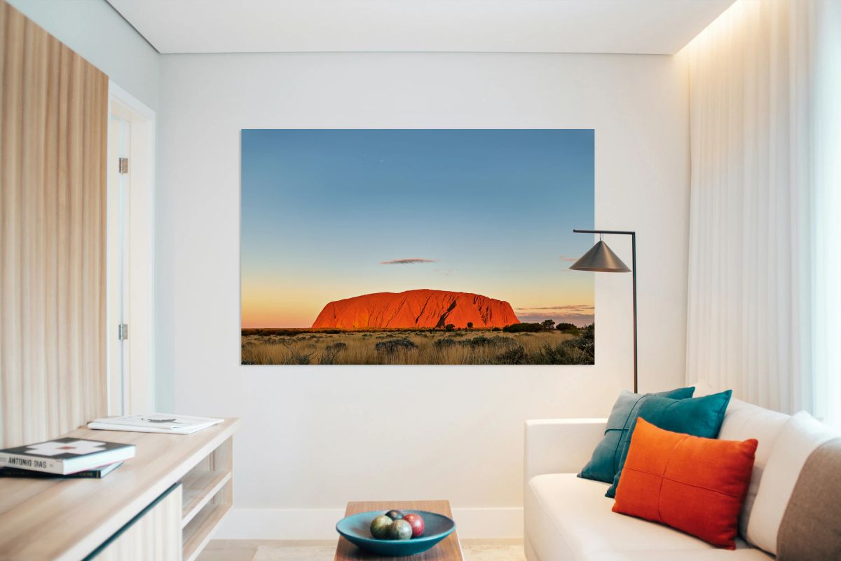 Uluru glows orange at sunset under a clear blue sky.